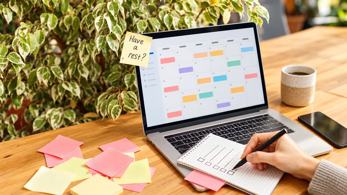A laptop showing a monthly planning grid beside a notebook and colourful sticky notes